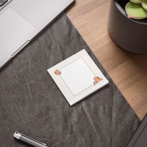 May include: A stack of square sticky notes with a brown border and cartoon bear illustrations in the corners. The notes are on a dark gray surface, near a laptop and a potted plant.
