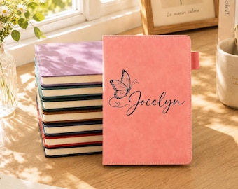 Può includere: Una pila di quaderni rettangolari con copertine a tinta unita nei toni del viola, blu, verde, marrone, rosso e nero. Un quaderno rosa è posizionato verticalmente davanti, con una grafica a farfalla nera e il nome Jocelyn in corsivo.