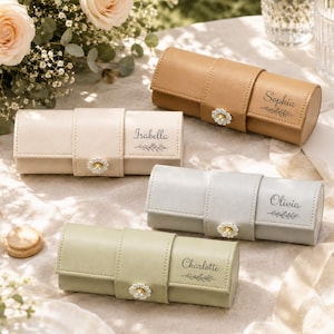 Może przedstawiać: Cztery cylindryczne etui na biżuterię w kolorach brązowym, kremowym, jasnoszarym i szałwiowym. Każde etui posiada środkowy pasek z białym zapięciem w kształcie stokrotki oraz jest spersonalizowane imionami Isabella, Sophia, Olivia lub Charlotte.