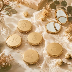 May include: A collection of round, gold-toned metal compact mirrors, each featuring a single engraved cursive letter on the lid. One mirror is shown open, revealing two reflective surfaces, while the others are closed, displaying the letters K, M, E, A, and S.