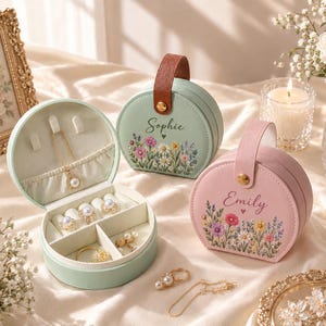 May include: Two round jewelry cases in sage green and soft pink, featuring floral prints and the names Sophie and Emily. The open green case reveals a necklace, rings, and compartments. Each case has a brown or pink handle with a gold snap.