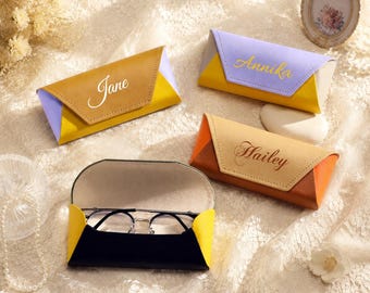 Può includere: Quattro custodie per occhiali personalizzate in marrone, arancione, giallo e viola. Ogni custodia ha un colore a contrasto sui lati ed è incisa con un nome. Una custodia è aperta, rivelando un paio di occhiali.
