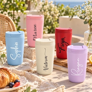 May include: Five insulated travel mugs in light blue, pink, cream, red, and lavender, each featuring a matching lid and a personalised name in a cursive script font. The names displayed on the mugs are Sophia, Maren, Victoria, Freya, and Reagan.
