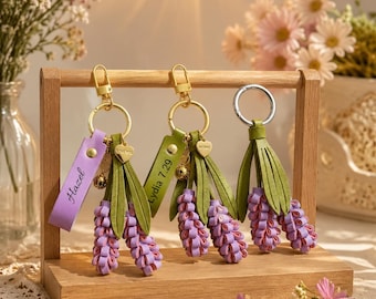 Può includere: Tre portachiavi con grappoli di fiori color lavanda e foglie verdi, fissati ad anelli dorati. Due portachiavi includono cinturini in pelle personalizzati con i nomi Hazel e Lydia 7.29, oltre a piccoli ciondoli a forma di cuore.