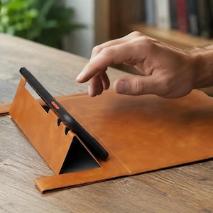 May include: A tan leather tablet stand with a triangular folding design that supports a black tablet in a landscape orientation. The stand features a smooth surface and a slot to hold the device securely in place while resting on a flat surface.