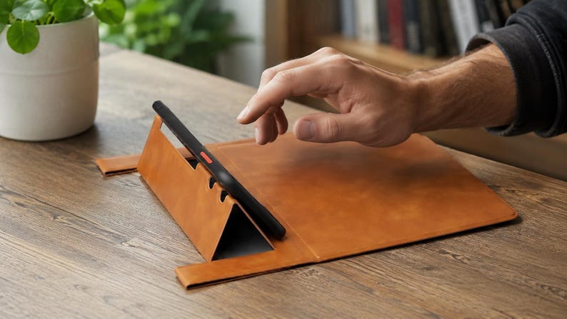 May include: A tan leather tablet stand with a triangular folding design that supports a black tablet in a landscape orientation. The stand features a smooth surface and a slot to hold the device securely in place while resting on a flat surface.