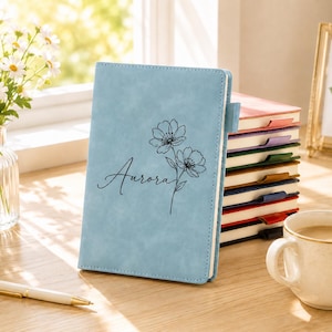 Op de afbeelding: Een lichtblauw notitieboek met een matte afwerking, voorzien van een zwarte lijntekening van twee bloemen en de naam Aurora in cursief schrift. Het notitieboek heeft een penlus en wordt getoond naast een stapel vergelijkbare notitieboeken.