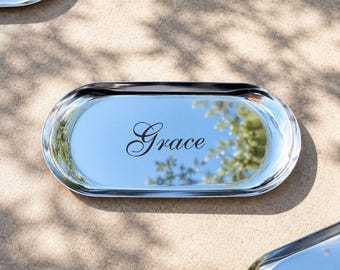 Può includere: Vassoio ovale a specchio con il nome "Grace" in elegante scrittura. La superficie riflettente riflette il cielo e gli alberi sovrastanti. Ulteriori vassoi sono visibili sullo sfondo, suggerendo un set. I vassoi sono in metallo lucido.
