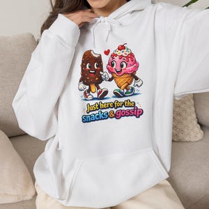 Op de afbeelding: Een witte hoodie met een afbeelding van een met chocolade bedekt ijsje en een roze bolletje ijs in een wafelhoorntje die elkaars hand vasthouden. Onder de figuren staat de tekst Just here for the snacks & gossip in kleurrijke letters.