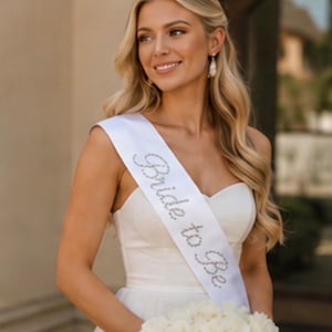 Könnte beinhalten: Eine weiße Satinschärpe mit dem Schriftzug Bride to Be aus silbernen Strasssteinen. Die Schärpe wird diagonal über den Oberkörper von der Schulter bis zur Hüfte getragen und ist für Feierlichkeiten rund um bevorstehende Hochzeiten gedacht.