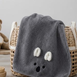 Peut inclure: Une serviette en peluche gris foncé présentant un motif de visage de koala avec deux oreilles duveteuses blanches et un nez noir. La serviette est drapée sur un panier en osier tressé. Le tissu a un aspect doux et texturé avec une bordure finie.