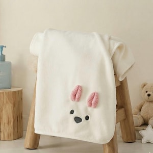 Peut inclure: Une serviette blanc cassé douce ornée d'un visage d'animal brodé avec deux petites oreilles roses texturées et des yeux et un nez sombres. La serviette est drapée sur un tabouret en bois, mettant en valeur une texture en tissu pelucheux.