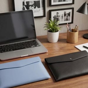 Peut inclure: Un espace de travail moderne avec un ordinateur portable, deux pochettes en cuir, une lampe de bureau et une petite plante en pot. Une pochette est bleu clair, l'autre est noire. Le bureau est en bois.