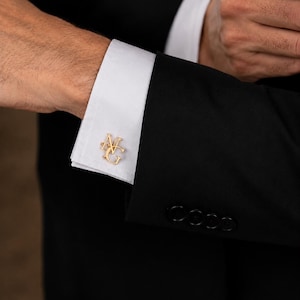 Peut inclure: Boutons de manchette monogrammés dorés avec les initiales "NG" sur un poignet de chemise blanche. La manchette est associée à une veste de costume noire. Accessoire classique pour les tenues formelles.