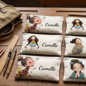 Peut inclure: Trousses à crayons en toile avec des illustrations de filles, chacune portant le nom "Camille". Un cartable en toile, des crayons, des stylos et une règle sont également sur une surface en bois. Les trousses présentent différents motifs de filles.