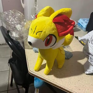 May include: A plush toy of a yellow fox-like creature with red accents on its ears and a white face. The toy has large red eyes and is standing on a light-colored surface. The plush is soft and cuddly, perfect for fans of the character.