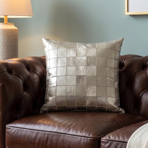 May include: A silver square patterned decorative pillow rests on a brown leather sofa. A cream-colored throw blanket with silver stripes is draped over the sofa. A lamp with a white shade and a small wooden side table are also visible.