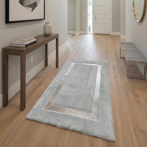 Puede incluir: Un pasillo moderno con suelo de madera clara, una alfombra azul claro con borde plateado y una consola de madera. Una obra de arte abstracta enmarcada cuelga de la pared y un espejo redondo está a la derecha.