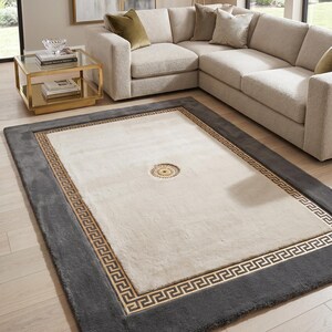 May include: A rectangular area rug with a cream-colored center and a dark gray border. The border features a Greek key design in a lighter brown color. The rug is placed in a living room setting with a sectional sofa and a glass-topped coffee table.