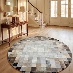 Puede incluir: Una alfombra circular grande compuesta por un mosaico de piezas cuadradas de piel en varios tonos de gris, beige, blanco, marrón y negro. La alfombra presenta un patrón de cuadrícula con una textura lisa de pelo corto sobre el suelo de madera clara.