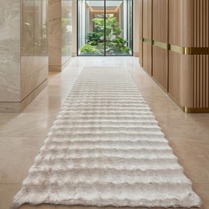 Puede incluir: Una alfombra larga y texturizada de color crema se encuentra sobre un suelo de piedra pulida en un pasillo. El pasillo presenta paneles de madera clara, paredes de mármol y una puerta de vidrio al final, que revela un pequeño jardín.