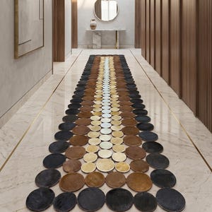 May include: A hallway runner rug composed of circular, textured elements in a gradient of colors: black, brown, tan, and white. The rug is centered on a marble floor, with wooden paneling on the walls and a decorative light fixture.