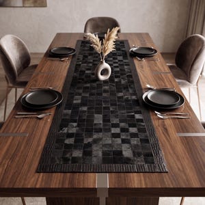 May include: A rectangular wooden dining table with a dark brown finish and metallic accents. A black checkered table runner sits in the center, topped with a small white vase containing dried pampas grass. Four place settings include black plates.
