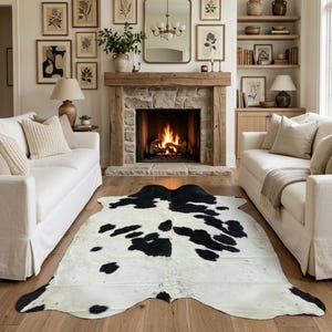 Peut inclure: Un tapis en peau de vache noir et blanc de forme organique irrégulière, présentant un motif naturel de taches sombres sur un fond clair, posé sur un parquet en bois clair entre deux canapés blancs dans un salon.