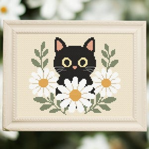 Puede incluir: Un bordado de punto de cruz en un marco de color claro que presenta un gato negro con grandes ojos amarillos rodeado de margaritas blancas con centros amarillos y hojas verdes sobre un fondo de tela color crema.