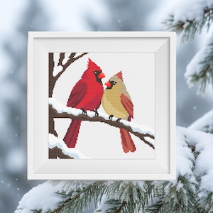 May include: A square white frame containing a cross-stitch pattern of two northern cardinal birds perched on a brown tree branch covered in white snow. The bird on the left is bright red with a black face mask, and the bird on the right is tan.