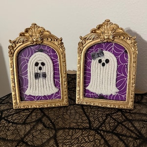 May include: Two decorative framed art pieces featuring white rope ghosts on a purple background with a white spiderweb pattern. Each ghost has black felt eyes and a mouth, with one wearing a black bow tie and the other a black hair bow.