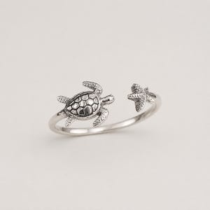 Op de afbeelding: Een zilverkleurige open ring met een gedetailleerde zeeschildpad aan het ene uiteinde en een gestructureerde zeester aan het andere. Het metaal heeft een geoxideerde afwerking in de verdiepte delen om het schildpatroon en de textuur te benadrukken.