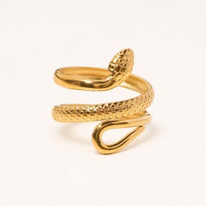 May include: A gold-coloured snake ring with a textured, scale-like pattern. The ring wraps around the finger in a spiral design, with the snake's head raised at the top. The ring is made of a shiny, metallic material.