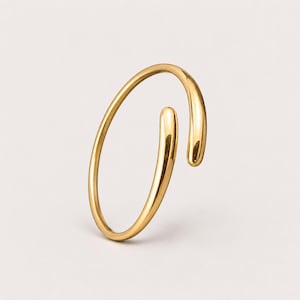 Puede incluir: Una pulsera abierta de tono dorado pulido con un diseño de alambre liso y redondeado. La pulsera tiene una forma circular con dos extremos redondeados y cónicos que no se unen, creando un espacio en la estructura de la banda metálica.