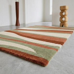 Könnte beinhalten: Ein rechteckiger Teppich mit einem Muster aus breiten, wellenförmigen Streifen in den Farben Terrakotta-Orange, Salbeigrün und Creme. Der Teppich hat eine dicke, getuftete Florstruktur und liegt auf einem glatten, hellgrauen Betonboden.