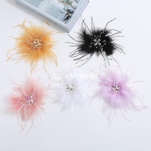 Op de afbeelding: Vijf decoratieve veren bloem broches in goud, zwart, roze, wit en lavendel. Elk stuk heeft een centraal cluster van heldere en gekleurde kristallen omringd door zachte, pluizige veren. De tekst Lacemoonlit is zichtbaar.
