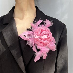 Op de afbeelding: Een roze stoffen rozenbroche met gelaagde bloemblaadjes en zachte roze veren, geaccentueerd met een klein stukje zwarte kant. De tekst LaceMoonlit is zichtbaar in het midden van de bloem. De broche is op een zwarte blazer gespeld.
