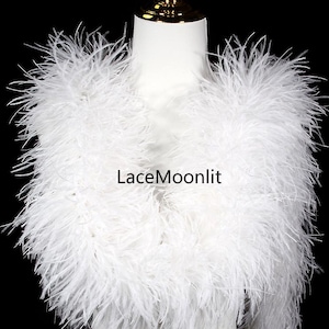 May include: A white feather shrug featuring long, wispy plumes draped around the neck and shoulders on a mannequin. The text LaceMoonlit is centered in black lettering over the white feathers, creating a soft, textured appearance.