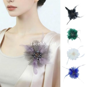 Op de afbeelding: Een bloemvormige broche met gelaagde stoffen bloemblaadjes en zachte veren in de kleuren paars, grijs, groen, wit en blauw. Het midden is versierd met donkere kralen. De tekst LaceMoonlit is zichtbaar op de afbeelding.