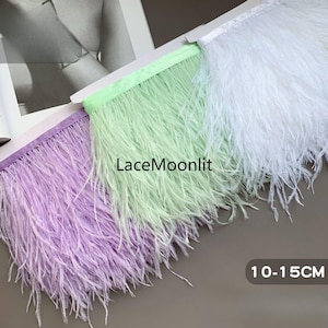 Puede incluir: Tres tiras de adorno de plumas de avestruz en lavanda, verde menta y blanco, cada una unida a una cinta de tela a juego. El texto "LaceMoonlit" está centrado en la tira verde menta y una etiqueta indica una longitud de 10-15 cm para las plumas.