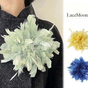 Op de afbeelding: Een saliegroene veren broche op een donkergrijs gestructureerd kledingstuk. Rechts worden twee extra veren broches getoond in geel en blauw. De tekst LaceMoonlit en LaceEmily is zichtbaar op de afbeelding.
