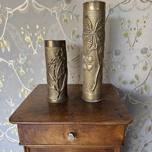 May include: Two cylindrical brass vases with textured surfaces. The shorter vase features a raised floral design with multiple petals. The taller vase displays a prominent embossed branch with several leaves. Both items have a metallic finish.