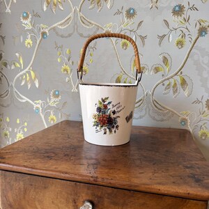 Può includere: Un secchiello in ceramica bianca di forma conica con un manico intrecciato marrone. La parte anteriore presenta un'illustrazione floreale colorata e il testo scritto a mano "Bonne fête Maman" in inchiostro nero. Il bordo ha una finitura marrone.