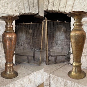 Peut inclure: Deux grands bougeoirs métalliques avec une base et un sommet évasés. Le bougeoir de gauche présente une finition aux tons cuivrés chauds, tandis que celui de droite a une finition aux tons laiton. Les deux ont un col fin et un corps arrondi.