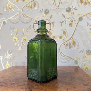 Könnte beinhalten: Eine quadratische Flasche aus smaragdgrünem Glas mit einem goldfarbenen Metalldrahtgriff am Flaschenhals. Die Flasche weist auf der Vorderseite und den Seiten geprägten Text auf, darunter die Wörter Distillerie De L'Etoile und Pellet France.