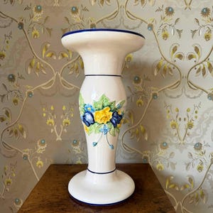 Peut inclure: Un vase sur pied en céramique blanche avec un haut et une base évasés, ornés de fines bordures bleues. Le corps central présente un motif floral en relief peint à la main, composé de roses jaunes et bleues entourées de feuilles vertes.