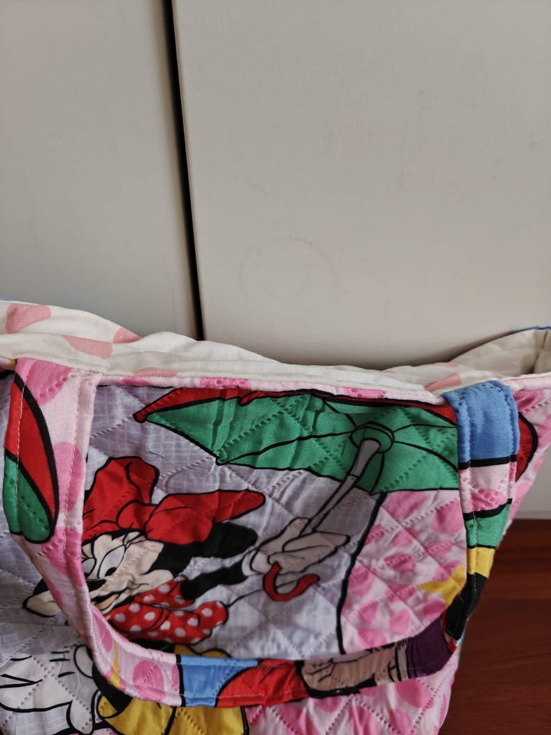 Może przedstawiać: Pikowana torba typu tote z nadrukiem Myszki Minnie. Torba ma r&oacute;żową, czerwoną i białą kolorystykę z rombowym wzorem. Uchwyty są niebieskie. Torba jest wykonana z tkaniny i jest przeznaczona do przenoszenia przedmiot&oacute;w.