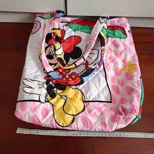Może przedstawiać: Pikowana torba typu tote z motywem Myszki Minnie. Torba jest gł&oacute;wnie r&oacute;żowa z białym pikowanym wzorem i dużym wizerunkiem Myszki Minnie. Uchwyty są wielokolorowe. Torba ma około 38 cm szerokości.