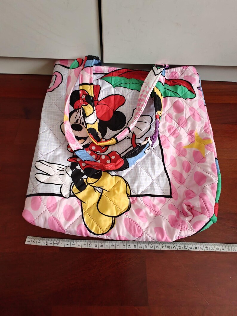 Może przedstawiać: Pikowana torba typu tote z motywem Myszki Minnie. Torba jest gł&oacute;wnie r&oacute;żowa z białym pikowanym wzorem i dużym wizerunkiem Myszki Minnie. Uchwyty są wielokolorowe. Torba ma około 38 cm szerokości.