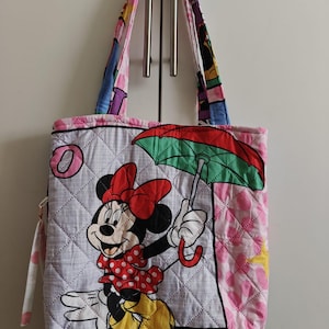 Może przedstawiać: Pikowana torba na zakupy z Minnie Mouse trzymającą parasol. Torba ma białe tło z rombowym wzorem i r&oacute;żowo-białą obw&oacute;dką w kropki. Uchwyty są wielokolorowe.